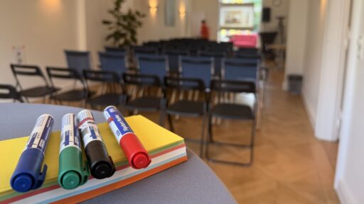 Stifte liegen auf einen Stapel bunten Papiers, im Hintergrund ein Seminarraum