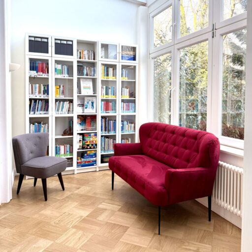 Ein rotes Sofa, ein kleiner Sessel und ein Bücherregal in der Wintergarten-Bibliothek der Löwenherz-Akademie