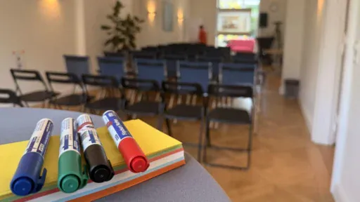 Stifte liegen auf einen Stapel bunten Papiers, im Hintergrund ein Seminarraum