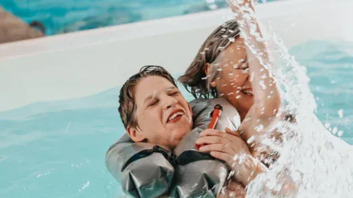 Eine ältere Frau planscht mit ihrem Enkel in einem Schwimmbad, der Wasser spritzt, beide lachen.