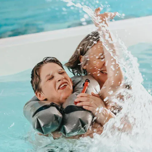 Eine ältere Frau planscht mit ihrem Enkel in einem Schwimmbad, der Wasser spritzt, beide lachen.