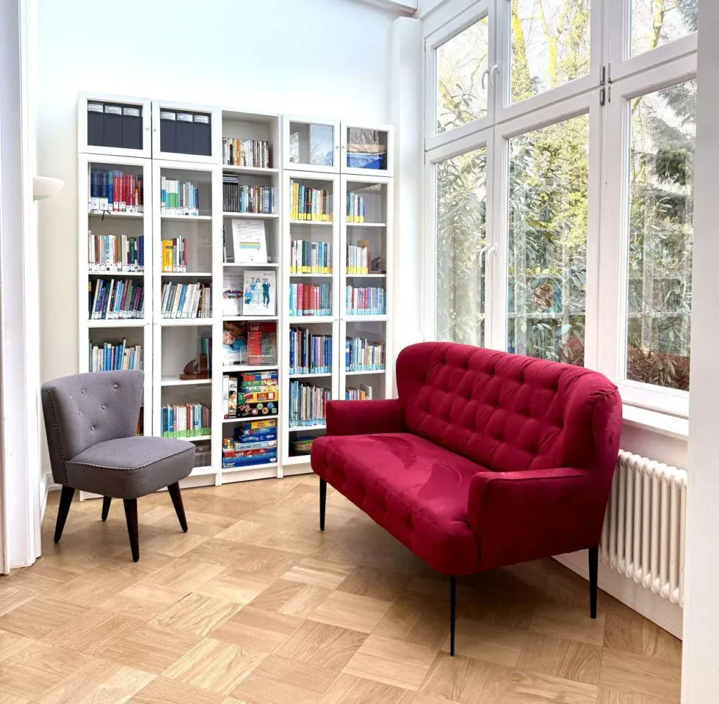Ein rotes Sofa, ein kleiner Sessel und ein Bücherregal in der Wintergarten-Bibliothek der Löwenherz-Akademie